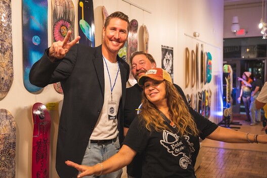 Gallery manager and friends smiling for camera at exhibit of skateboard art hung on picture hanging system